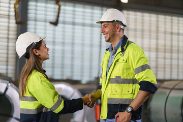 A man and a woman workers are standing in a factory, They wear a yellow and white safety helmet for safety.