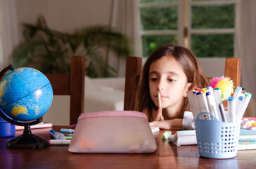 A young girl doing her homework on a wooden table surrounded by school supplies.