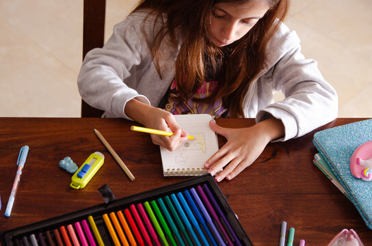 A young girl coloring a hand drawn draw on a wooden table, surrounded by school and art supplies.
 - Powered by Adobe