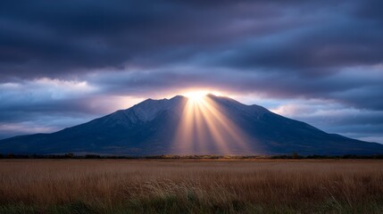 Obraz premium Majestic mountain peak illuminated by dramatic sunbeams piercing through dark cloudy skies