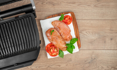 Two raw marinated chicken breasts rest on a wooden cutting board with basil leaves and tomato...