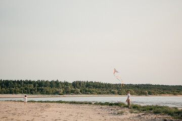 A father helps their daughter fly a colorful bird-shaped kite on a sandy river beach under a bright sky