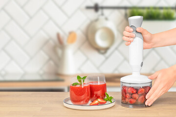 Hands operate a hand blender filled with strawberries next to a plate with two glasses of fresh strawberry smoothie garnished with mint on a wooden kitchen counter