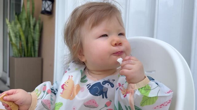Adorable baby girl in colorful bib sitting in a high chair, exploring food with curiosity while practicing baby led weaning with fruits, vegetables, and eggs during an outdoor mealtime at home