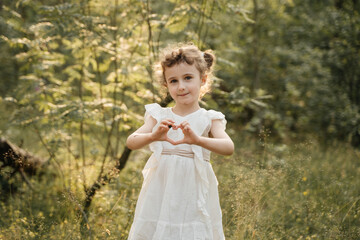 A cheerful child in a white dress, with curly hair styled in two buns, forms a heart shape with her...