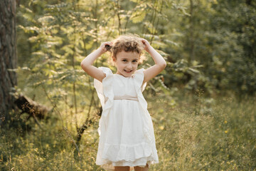 A cheerful child in a white dress, with curly hair styled in two buns, forms a heart shape with her...