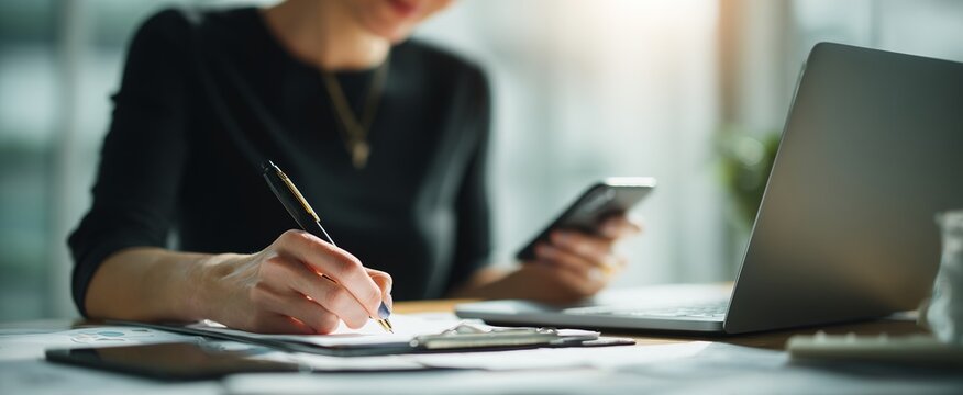 A businesswoman holds a paper and pen, working on a laptop computer at a wooden desk with books, papers, and a mobile phone in an office. This scene represents the business concept of working from hom