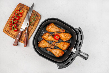 Five golden-brown chicken drumsticks with a cherry tomato and herbs rest in an air fryer basket beside a wooden cutting board with knives and fresh vine tomatoes