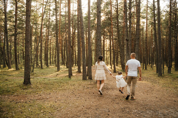 Fototapeta premium A happy family of mother, father, and daughter walks hand-in-hand away from the camera on a sun-dappled forest path.