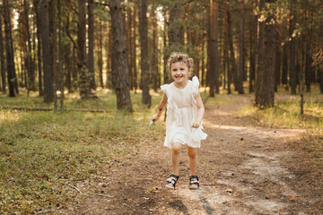 A cheerful child with curly hair and a white dress runs excitedly on a forest path, surrounded by...