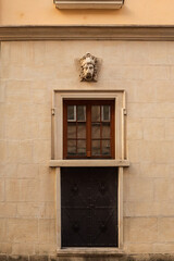Fototapeta premium Historic stone wall with small window, wooden frame, black metal door below, and decorative lion head sculpture above the frame in warm evening light