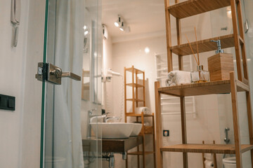 Bright modern bathroom interior with wooden shelving, rolled towel, fragrance diffuser, and soap...