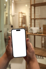 Hands holding a smartphone with blank white screen in modern bathroom. Wooden shelving, towel, and...