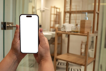 Hands holding a smartphone with blank white screen in modern bathroom. Wooden shelving, towel, and...