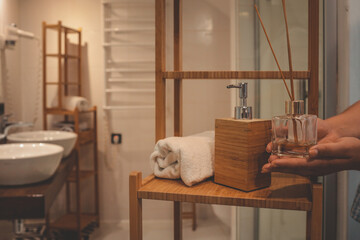 Hands arranging a reed diffuser on a wooden shelf in a cozy bathroom. White towel, soap dispenser,...