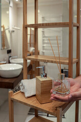 Hands arranging a reed diffuser on a wooden shelf in a cozy bathroom. White towel, soap dispenser,...