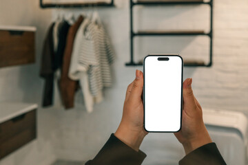 Person holding smartphone with blank white screen in modern loft-style closet with hanging clothes, shelves, and warm lighting against a white brick wall