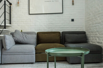 Modern living room with a small green metal coffee table in focus and a cozy sectional sofa in neutral tones against a white brick wall in the background