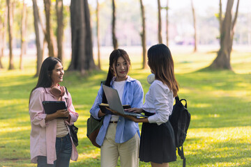 Group of Asian college students standing on green grass and discussing with laptop and books. Concept of teamwork, education, and outdoor learning in a natural environment.