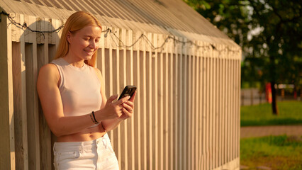 woman using smartphone outside in golden hour. smiling casually, she leans against wooden structure...