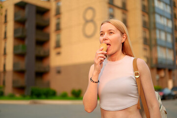 woman enjoying ice cream outdoors on sunny evening stroll near modern apartment complex. casual clothing, relaxed atmosphere, urban lifestyle, youthful vibe, suburban environment