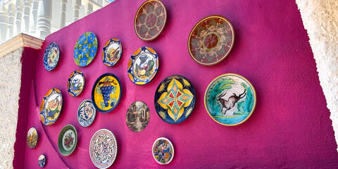 Colorful decorative plates arranged on a vibrant pink wall, showcasing intricate designs and patterns, creating an artistic and lively atmosphere in a modern exterior space
