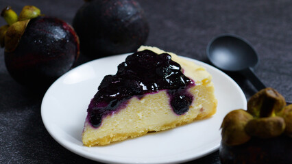 Creamy new york style cheesecake slice topped with sweet blueberry sauce, served on a white plate alongside whole mangosteens against a dark textured background
