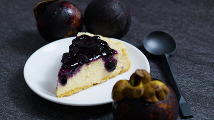 Creamy blueberry cheesecake slice served on a white plate with fresh mangosteens and a black spoon, creating a vibrant and tempting dessert presentation on a dark background