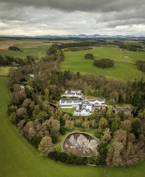 Aerial view of a grand white building nestled amidst lush green fields and dense woodland, framed by a circular feature, Peebles, Scotland, United Kingdom.
