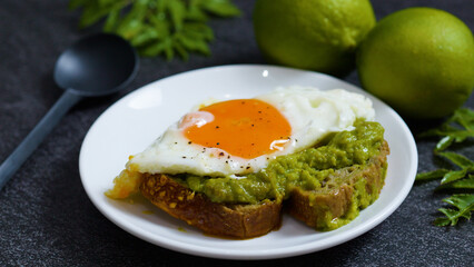 Whole grain bread topped with mashed avocado and fried egg seasoned with black pepper creates a vibrant and nutritious breakfast, accompanied by fresh limes on a dark background