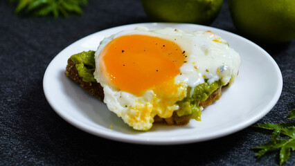 Whole grain toast topped with creamy mashed avocado, crispy fried egg sprinkled with black pepper on minimalist white ceramic plate, representing trendy healthy breakfast