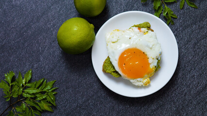 Top view of a vibrant and appetizing breakfast featuring fried egg on avocado toast, fresh limes, and green leaves on a dark background