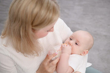 Mom holds newborn baby in arms with care, baby drinks baby formula from bottle, which is carefully...