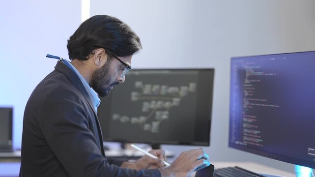 Focused Indian male AI developer coding a complex machine learning algorithm late at night. A dedicated data scientist working on a neural network at a multi-moni