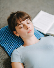 Fototapeta premium A young woman lies on a blue acupressure mat with closed eyes, appearing relaxed and meditative beside an open book.
