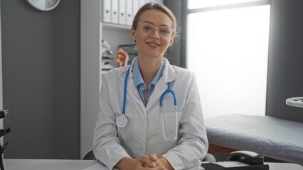 Female doctor sitting in a hospital room, smiling and wearing a white coat with stethoscope, embodying professionalism and trust in a clinical indoor setting.