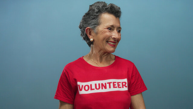 Woman smiling with grey hair in red volunteer shirt against blue background, expressing joy and community spirit, emphasizing older female engagement in social causes.
