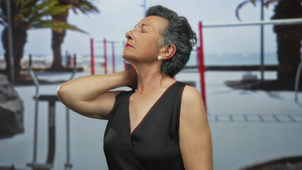Senior woman with grey hair wearing a sleeveless dress, touching her neck while standing thoughtfully on a seaside promenade with blurred beach and fitness equipment in the background.