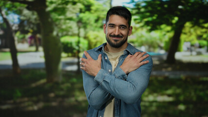 Obraz premium Young hispanic man smiling in a peaceful park setting, with arms crossed embracing himself under the lush green trees, exuding a sense of relaxation and contentment outdoors.