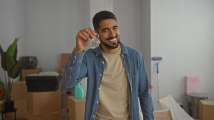 Young man holding house keys smiling in a new home interior surrounded by boxes indicating moving...