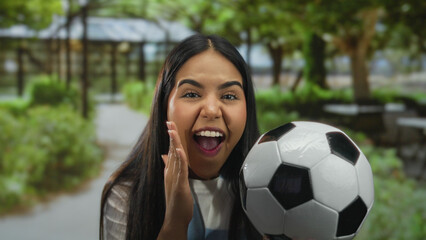 Woman cheering with soccer ball in lush green park setting, expressing excitement and joy in an outdoor environment, embracing soccer culture and passion.