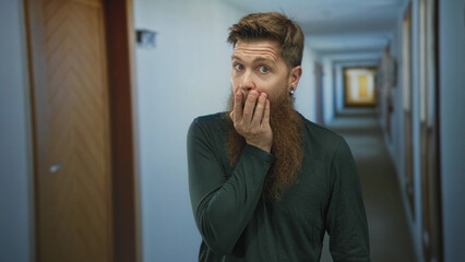 Young man with thick beard and finger to lips for silence in a hotel building corridor beside...