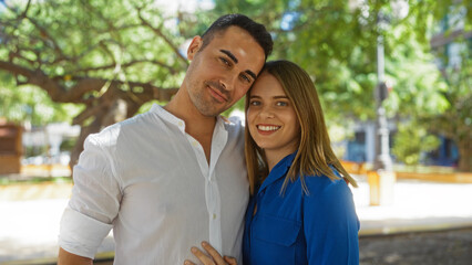 Obraz premium Man and woman embracing outdoors in a sunlit urban park, showcasing a hispanic couple enjoying a beautiful moment together in a city setting with trees around.