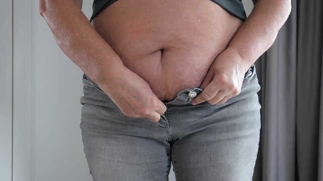 Overweight woman is struggling to button her tight jeans, highlighting the concept of weight gain and the need for a healthier lifestyle. Close up of the cellulite belly of an elderly diabetic female