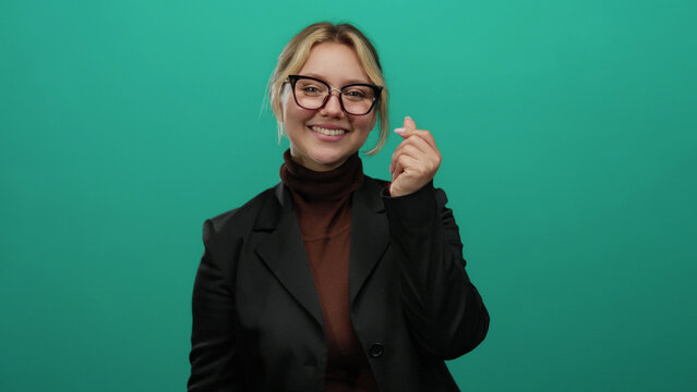 Woman making heart gesture with fingers smiling against a vibrant green background, showcasing confidence and positivity with her blonde hair and glasses.