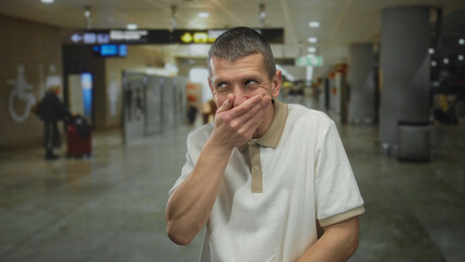 Caucasian man covering mouth in airport terminal with blurred background indicating surprise or...
