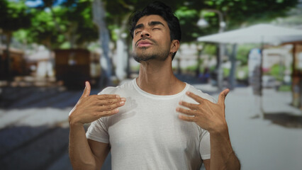 Young hispanic man fans himself with hands and wipes sweat from forehead on crowded street under...