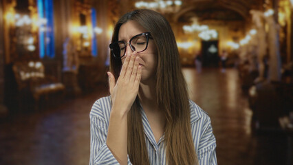 Woman pinches her nose with hand in ornate museum corridor lined with gilded columns under warm...