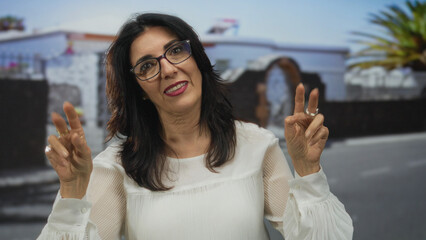 Woman wearing glasses and a white blouse with raised hands making a gesture on city street;...