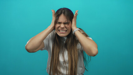 Hispanic woman in white shirt covers ears with hands against green wall in response to noise...